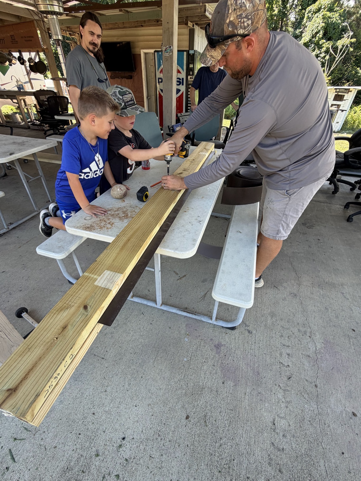 Adults and children working together on a lumber project at a picnic table under the camp shelter during a club workday.