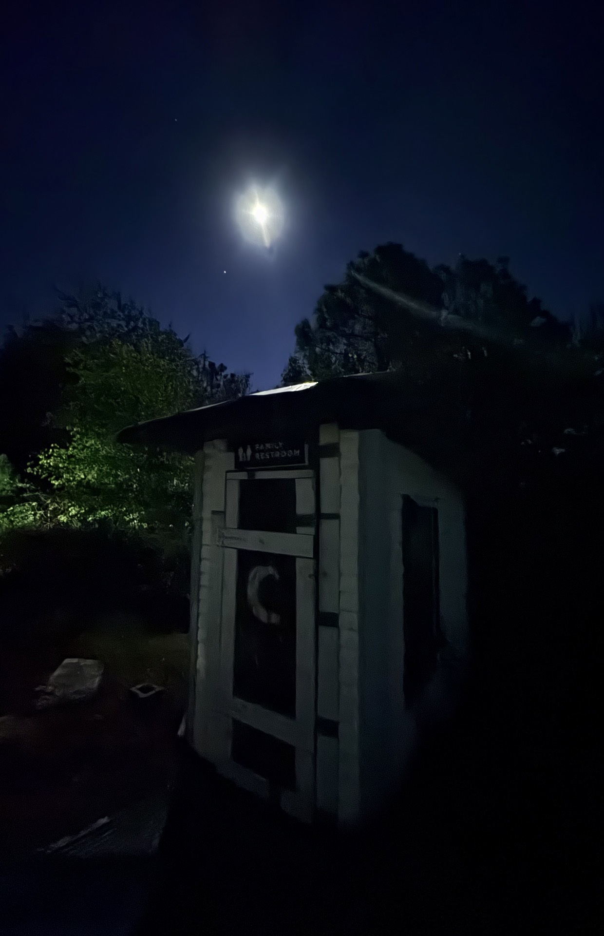 The camp outhouse beneath a bright moon, surrounded by trees at night.